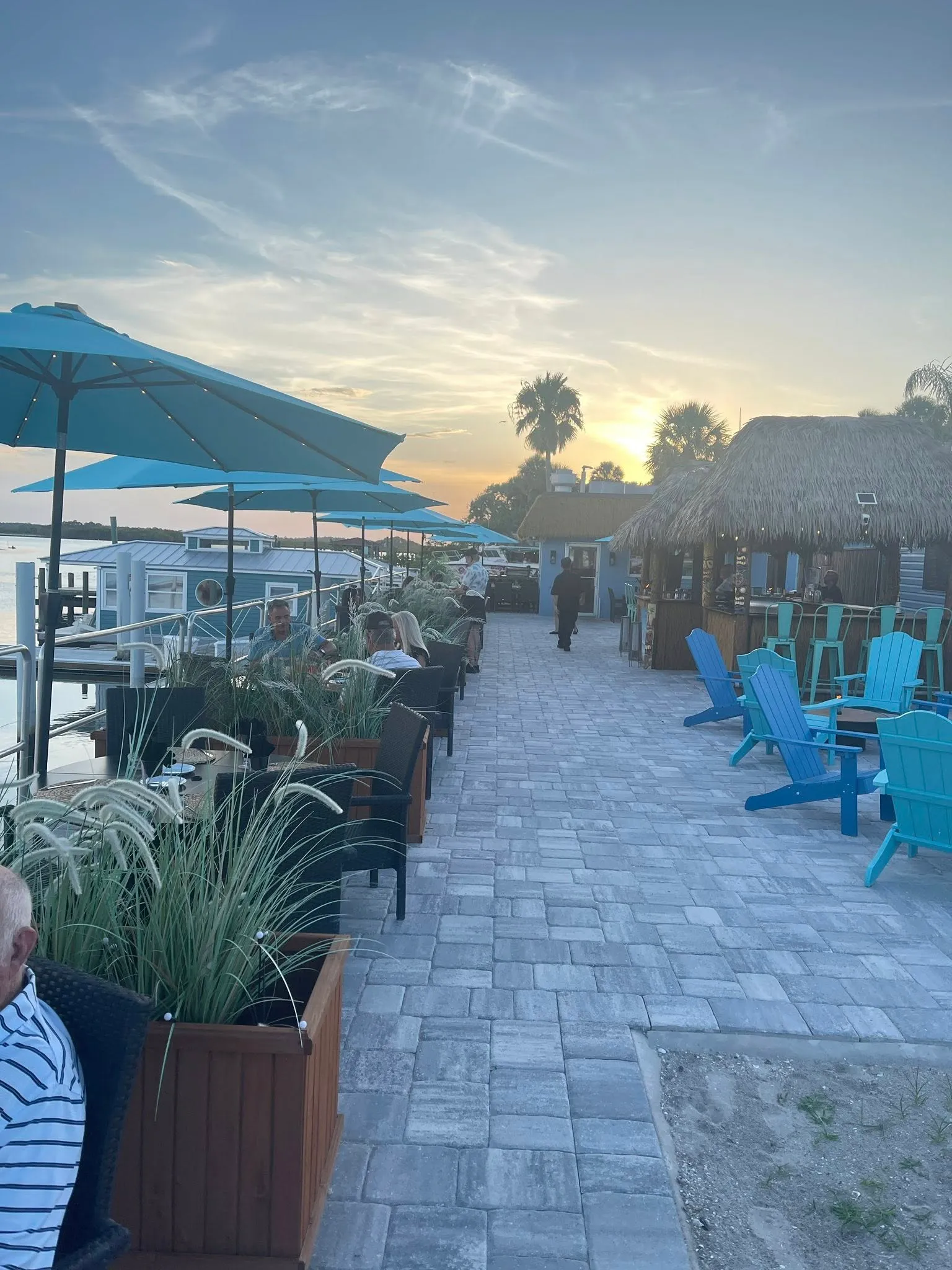 Outdoor waterfront dining area at HT's Sand Bar & Bistro with guests seated under blue umbrellas and a tiki-style bar.